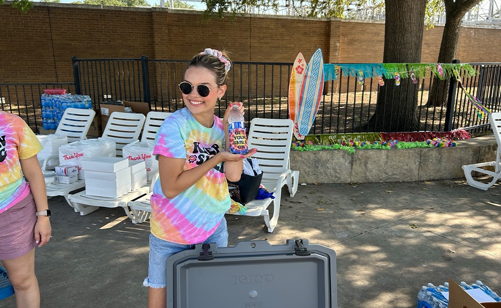 Woman in tie dye shirt gesturing to a drink with a tie dye coozie