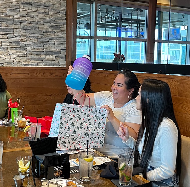 Woman at restaurant table opening a present with a large purple water bottle